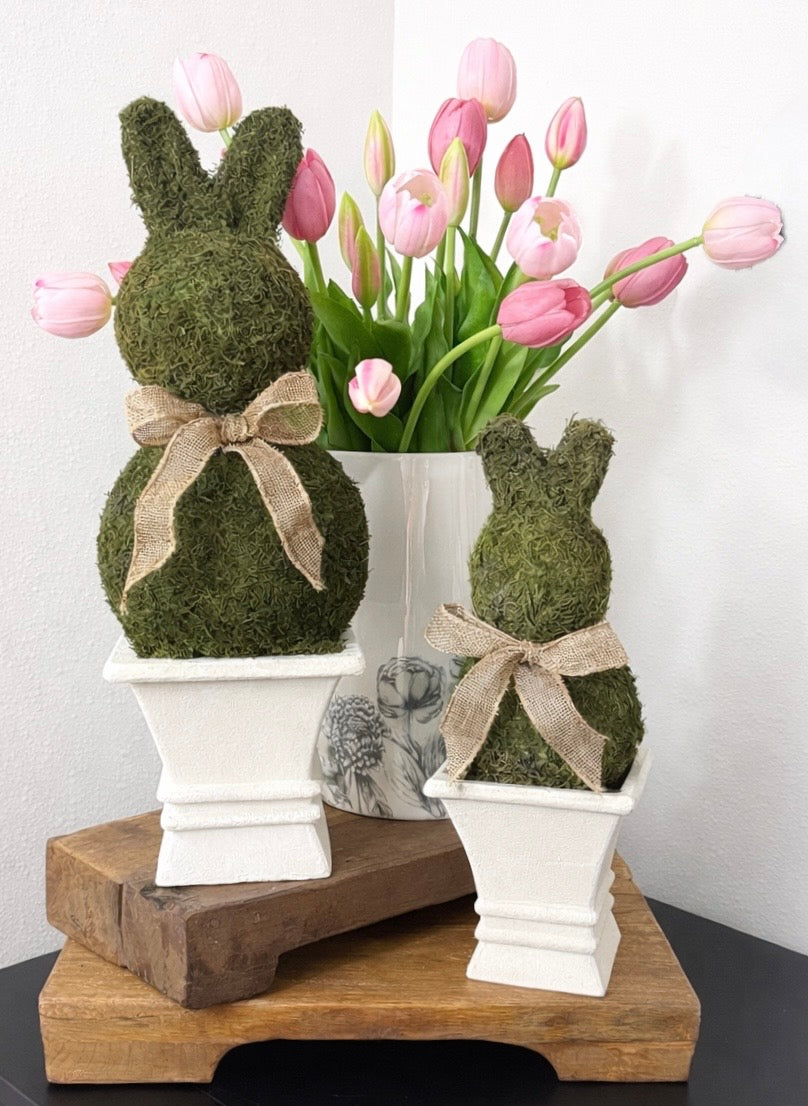 Two moss-covered bunny figures with ribbons in white pots, surrounded by pink tulips on a wooden stand.
