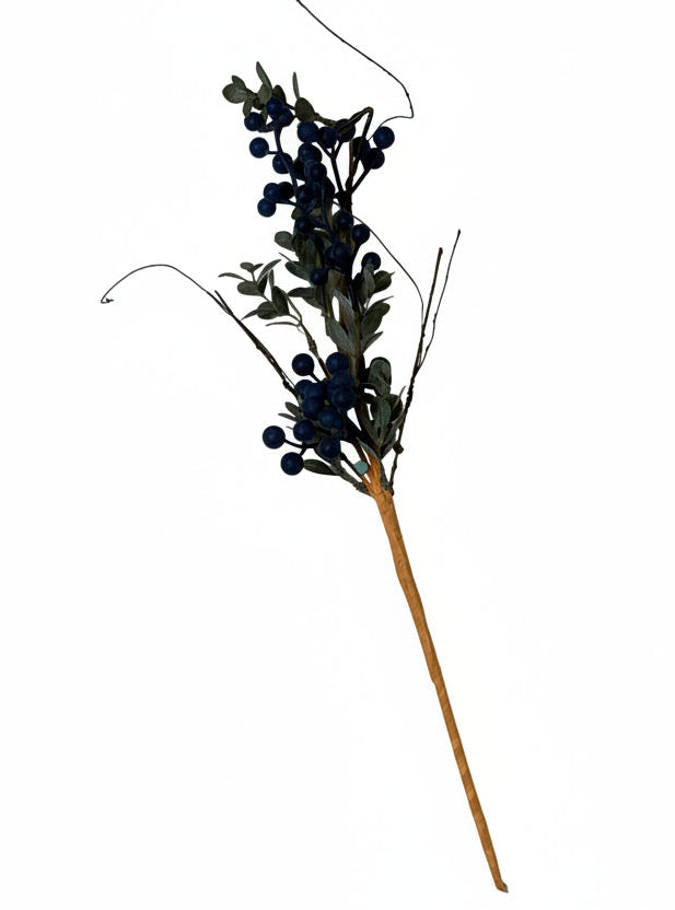 A single artificial floral stem featuring dark blue berries, muted green-gray leaves, and brown branches, displayed against a textured off-white wall.